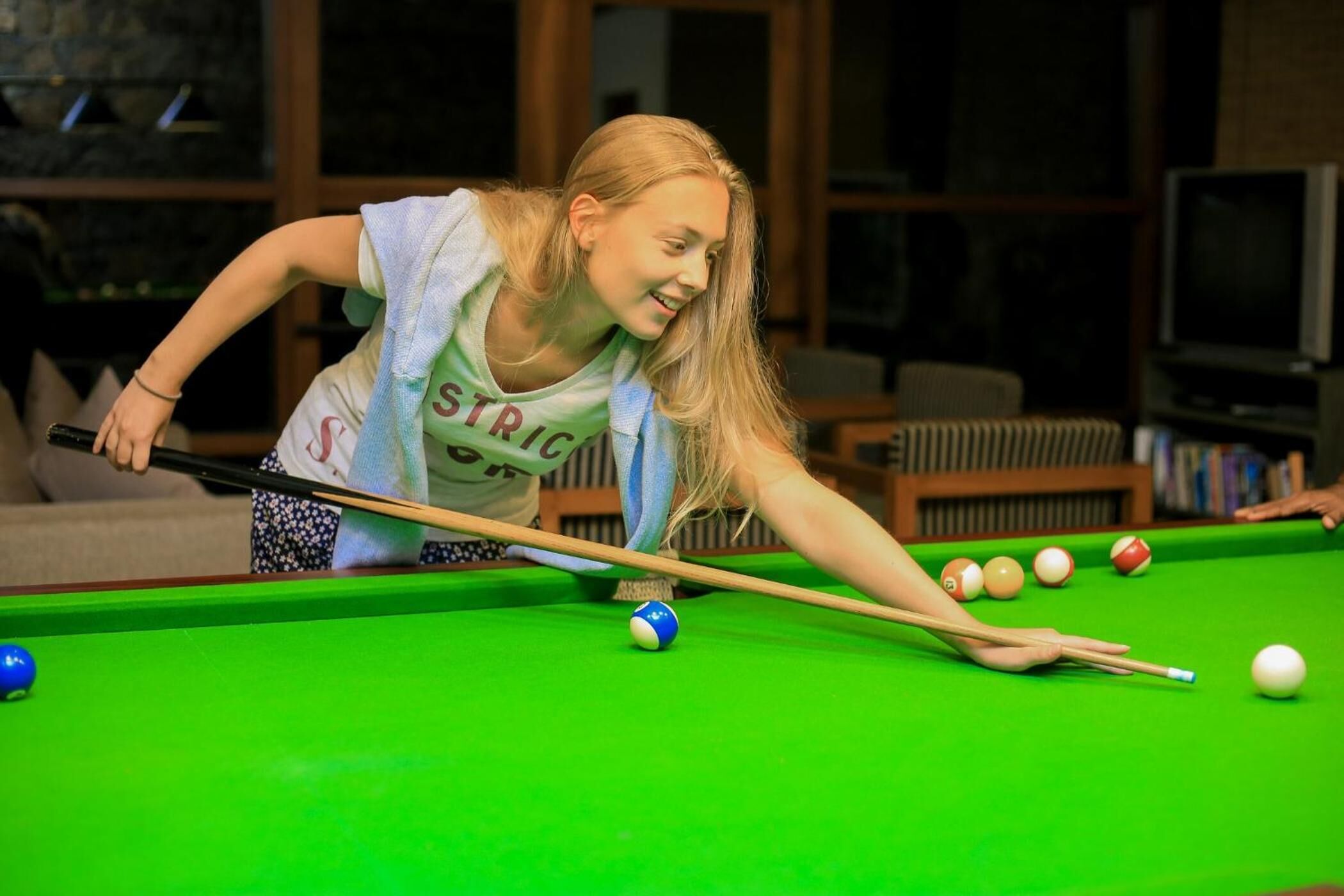 A person playing pool in a room with a green pool table, a sofa, and a television in the background.