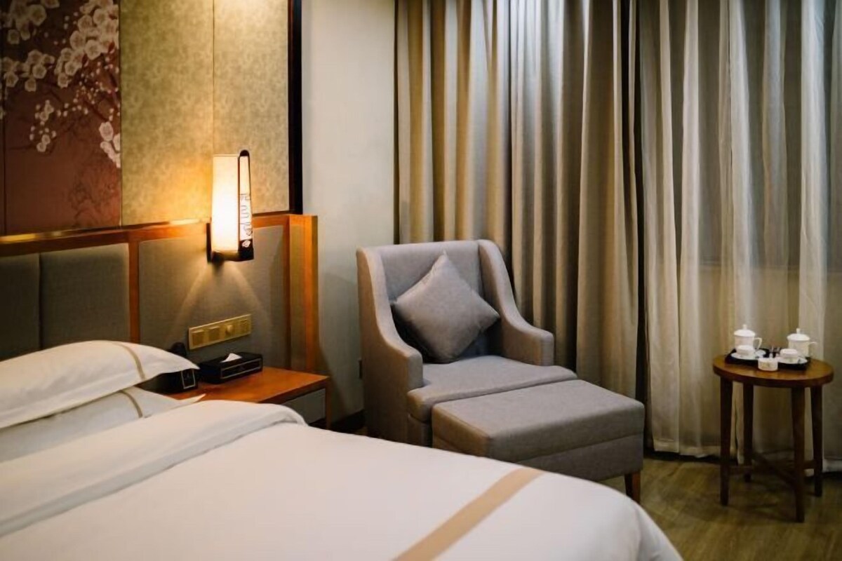 A hotel room with a bed, a chair, a bedside table with a lamp, and a small table with a teapot and cups.