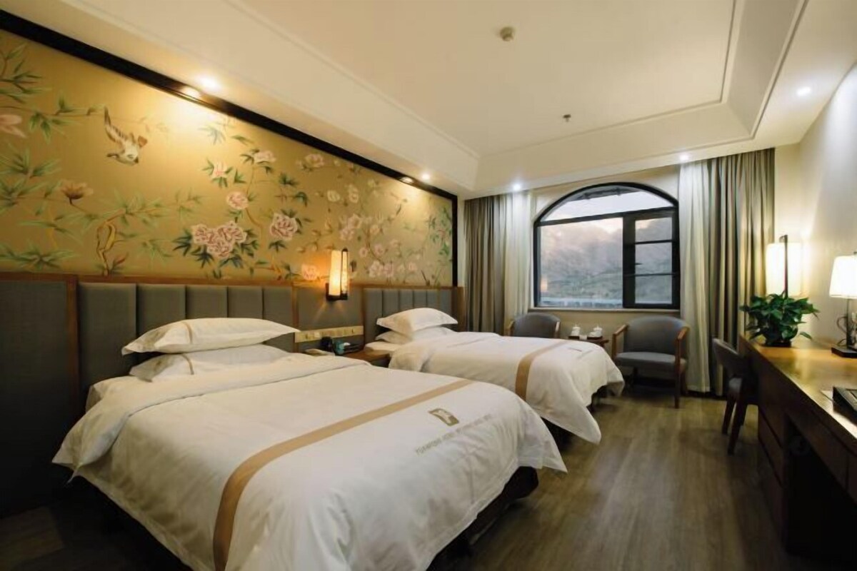 A hotel room with two beds, a desk, and a view of a mountain through a large window.