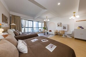 Quarto de hotel moderno com duas camas, área de estar com sofá e poltronas, escrivaninha e uma grande janela com vista para a cidade.