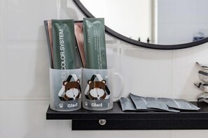 Casa de banho com parede revestida de azulejos brancos, bancada preta e espelho. Há duas chávenas com um desenho de urso e a palavra "Glad" nelas, juntamente com alguns artigos de higiene pessoal.