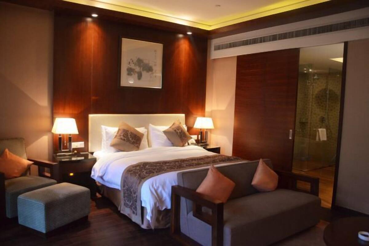 A modern hotel room with a large bed, two bedside tables with lamps, a sofa, and a glass shower area.