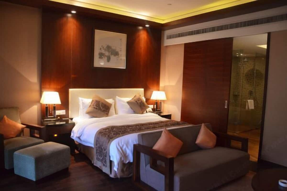 A hotel room with a large bed, two bedside tables with lamps, a sofa, and a glass door leading to a bathroom.