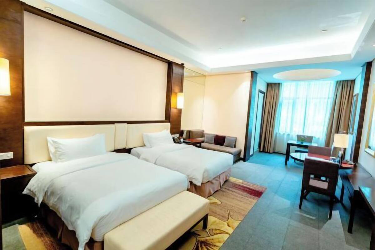 A hotel room with two beds, a desk, a chair, a sofa, and a large window with curtains.