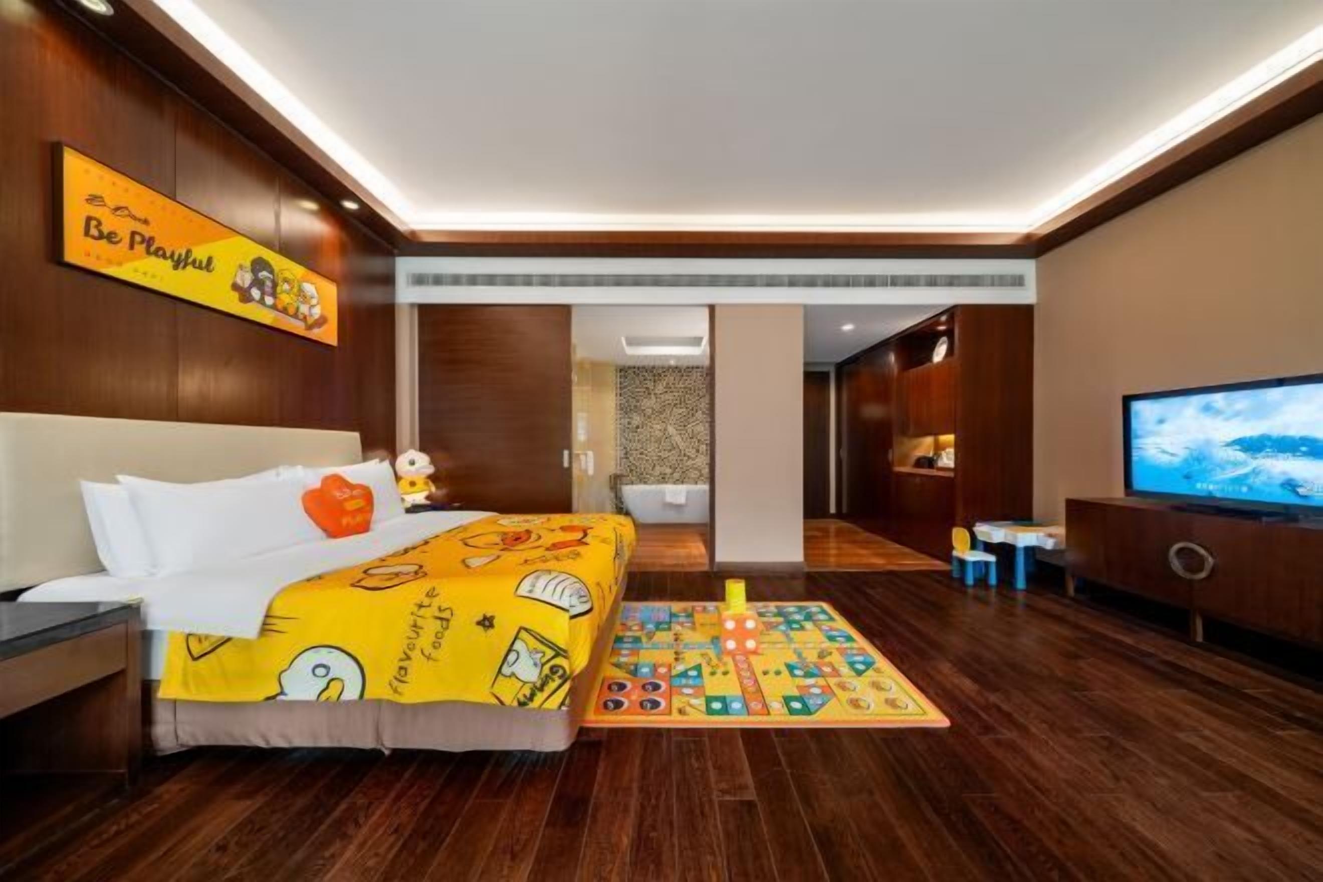Quarto de hotel com cama, tapete de brincadeira, televisão e piso de madeira.