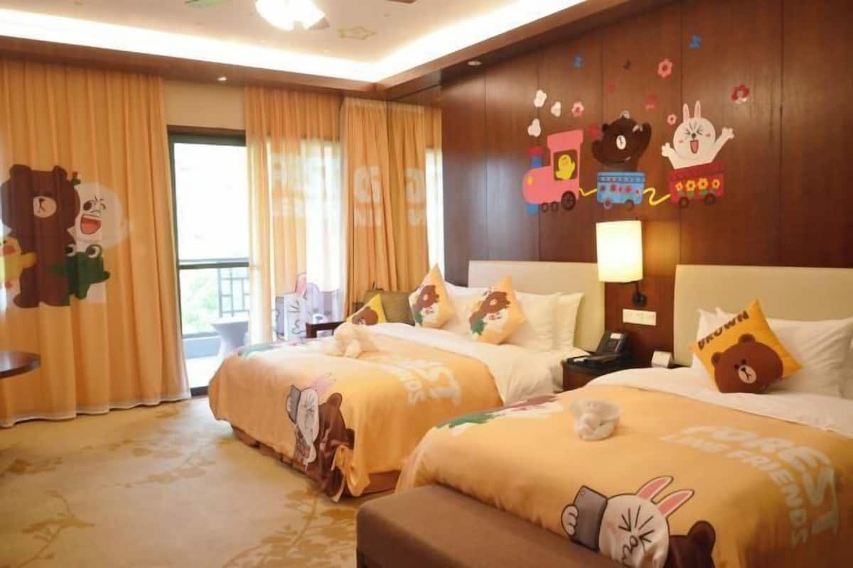 A hotel room with two beds, each adorned with cartoon character bedding. There is a large window with curtains, a bedside table with a lamp, and a wall with a cartoon train mural.
