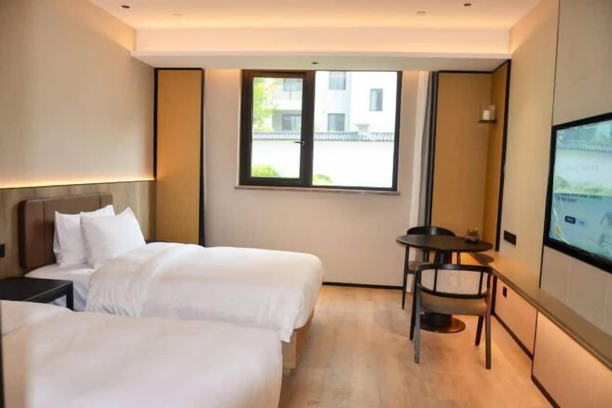 A hotel room with two beds, a small table, a chair, a TV, and a window with a view of buildings.