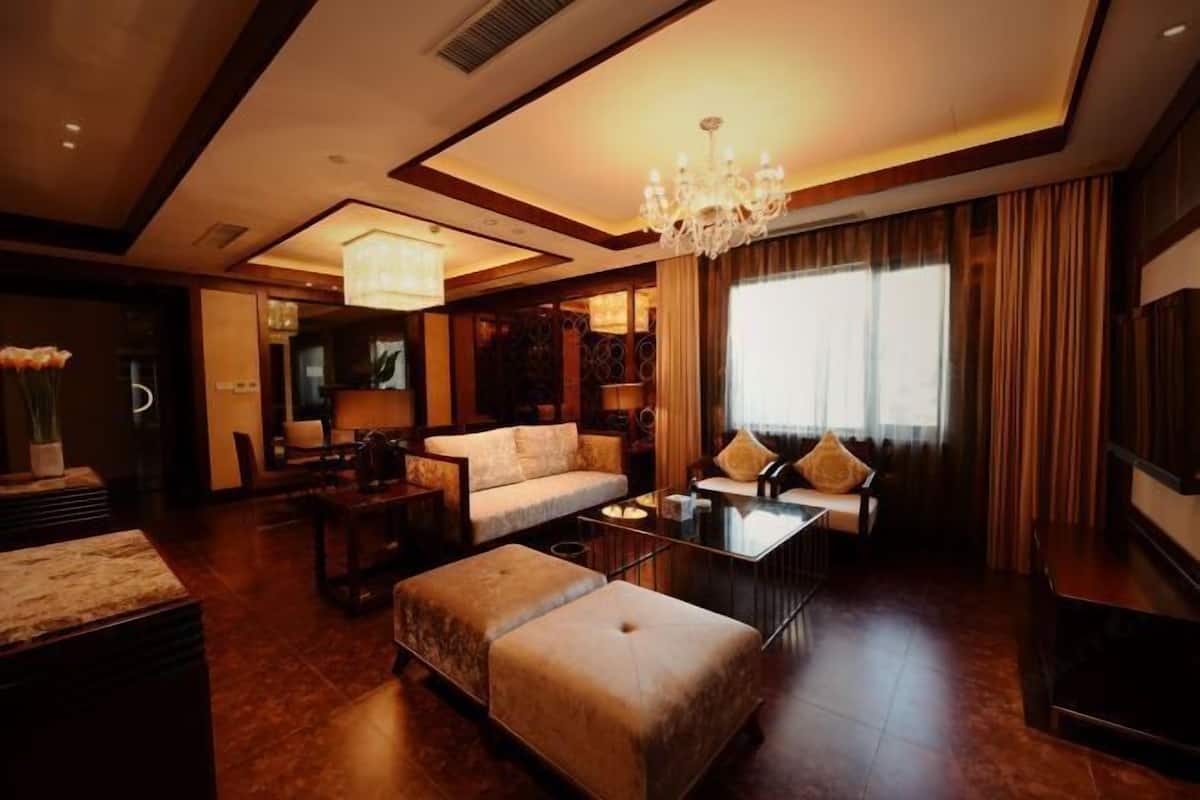 A spacious living room with a sofa, ottoman, and coffee table. There is a chandelier, a large window with curtains, and a television on the wall.