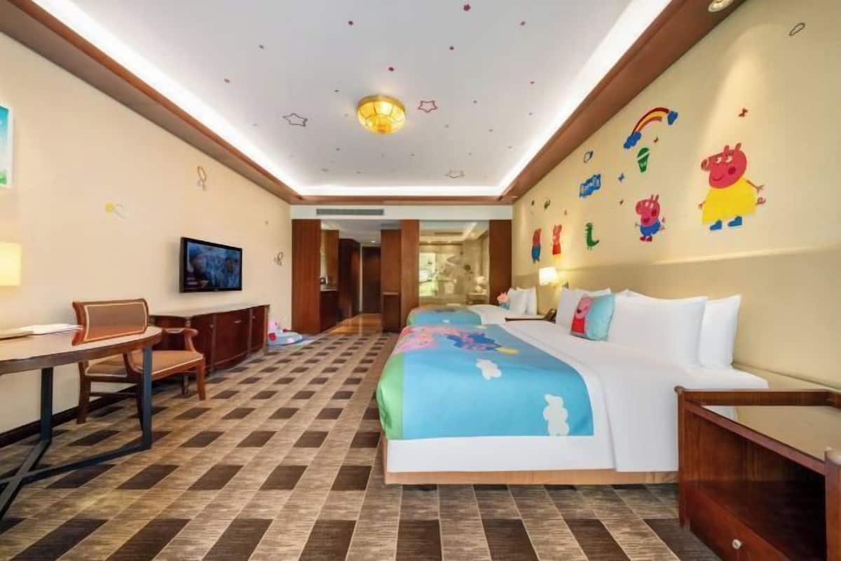 A hotel room with a checkered floor, a bed with colorful bedding, a desk with a chair, a television, and a wall with cartoon character decals.