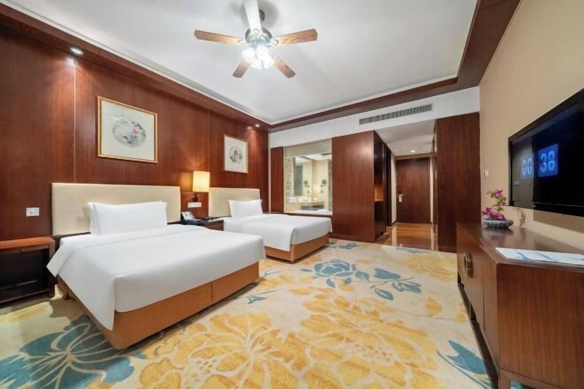 A hotel room with two beds, a ceiling fan, a TV, and a floral carpet.