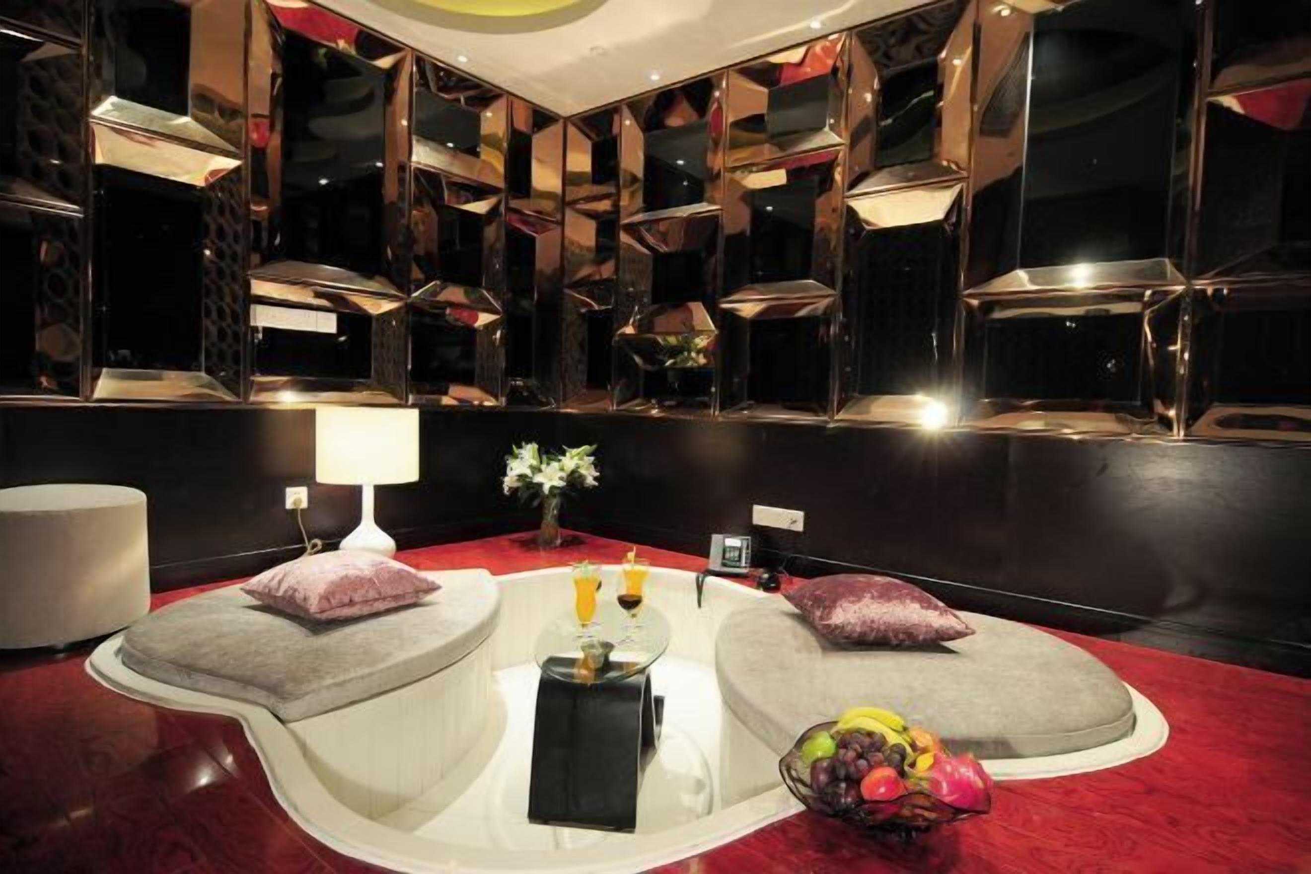 Une pièce moderne avec un tapis rouge, une table ronde, deux coins salon avec des fauteuils confortables, un mur miroir et un vase rempli de fleurs.