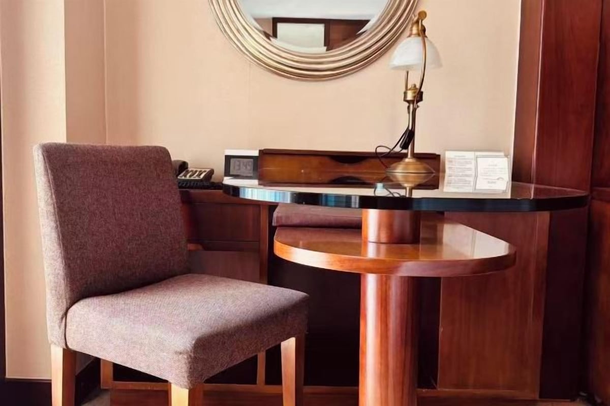 A hotel room with a round wooden table, a cushioned armchair, a small desk with a keyboard, a lamp, and a mirror on the wall.