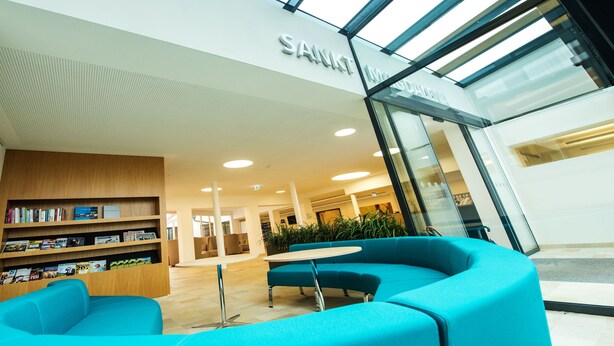 A modern lobby with a blue seating area, a wooden bookshelf, and a glass entrance to a room labeled "SANKT." - Sankt Magdalena (Linz)