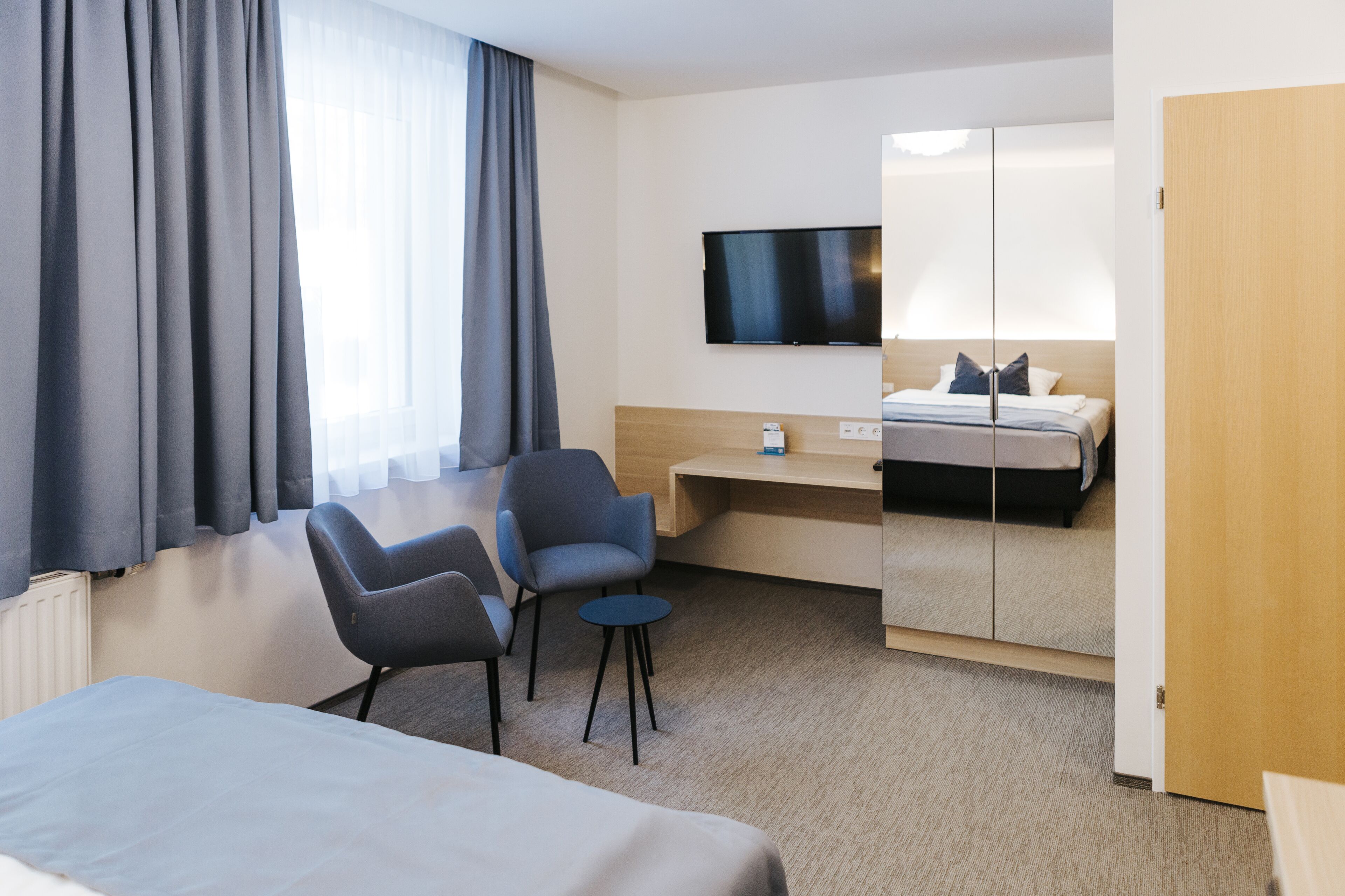 Quarto de hotel com cama, escrivaninha, duas cadeiras, televisão e uma janela grande com cortinas.