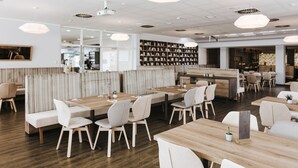 A modern restaurant with wooden tables and white chairs, a bookshelf in the background, and a counter area. - Sankt Magdalena (Linz)