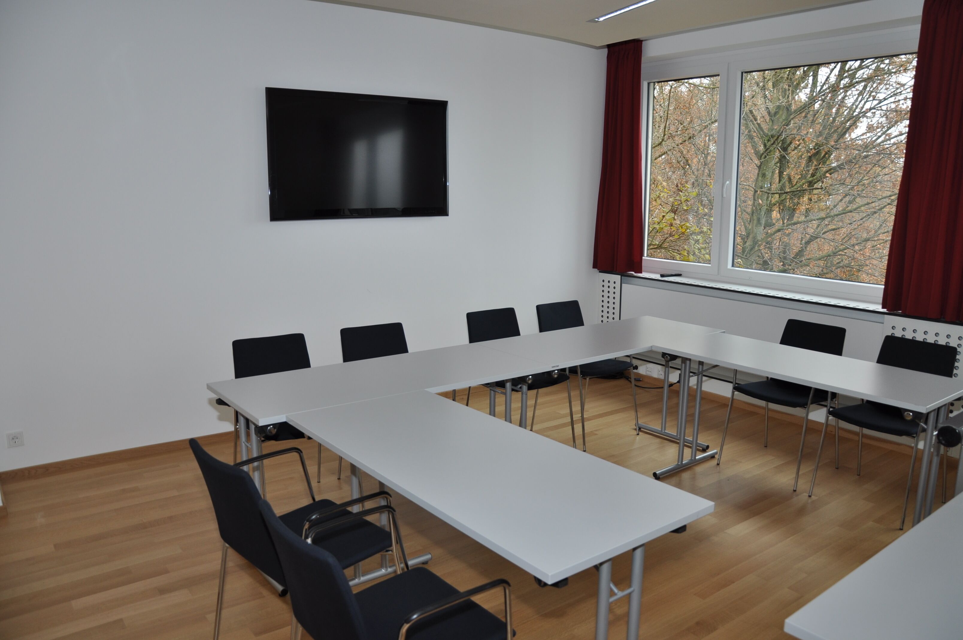 A conference room with a long table, black chairs, a flat-screen TV, and a window with curtains.