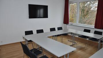A conference room with a long table, black chairs, a flat-screen TV, and a window with curtains.