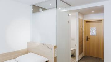 Una habitación de hotel moderna con un separador de vidrio, un cabecero de madera, una cama con ropa blanca y una puerta de madera. Se ve un inodoro a través del separador.