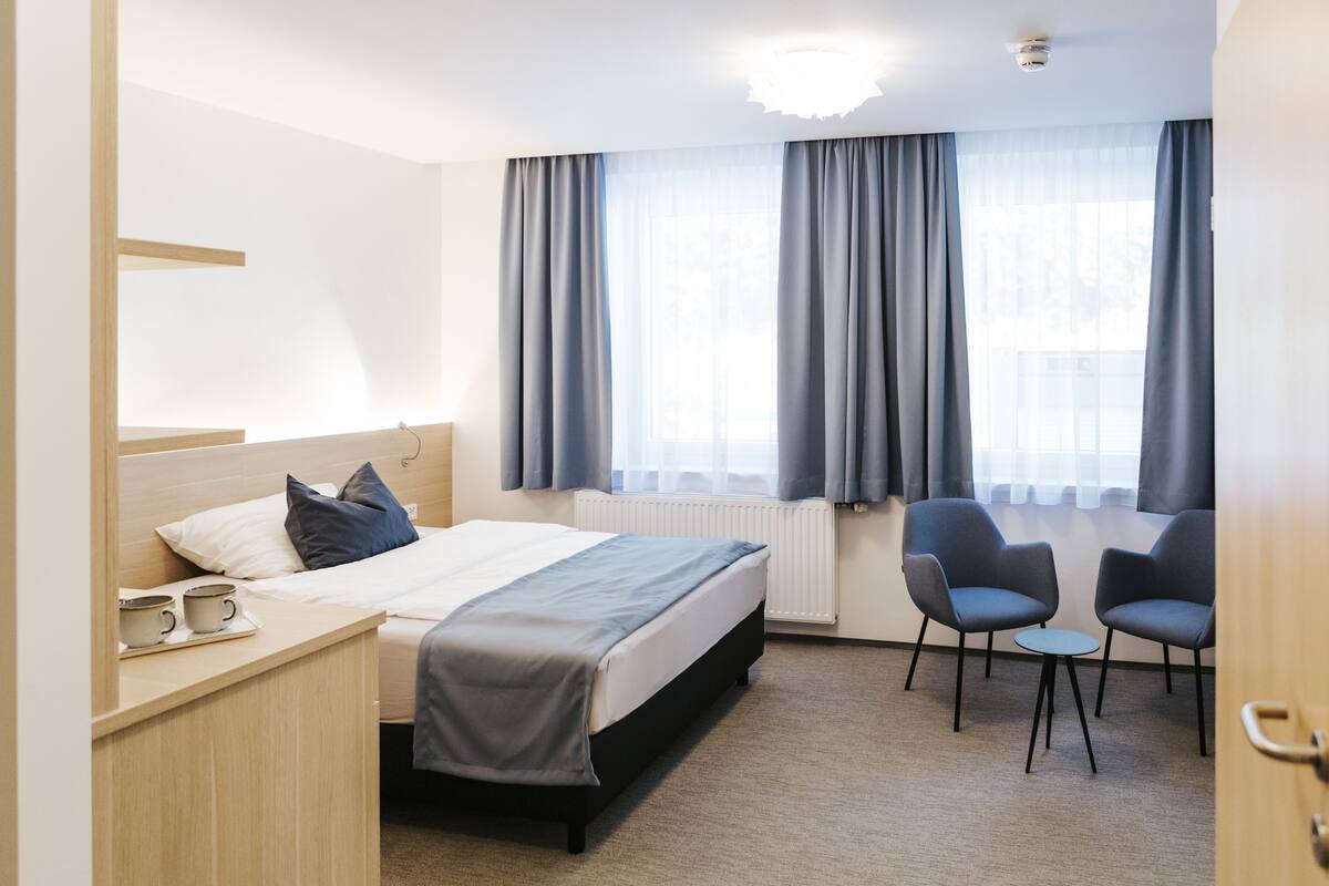 Habitación de hotel con cama, mesita de noche, dos sillas, una mesa pequeña y ventana con cortinas.