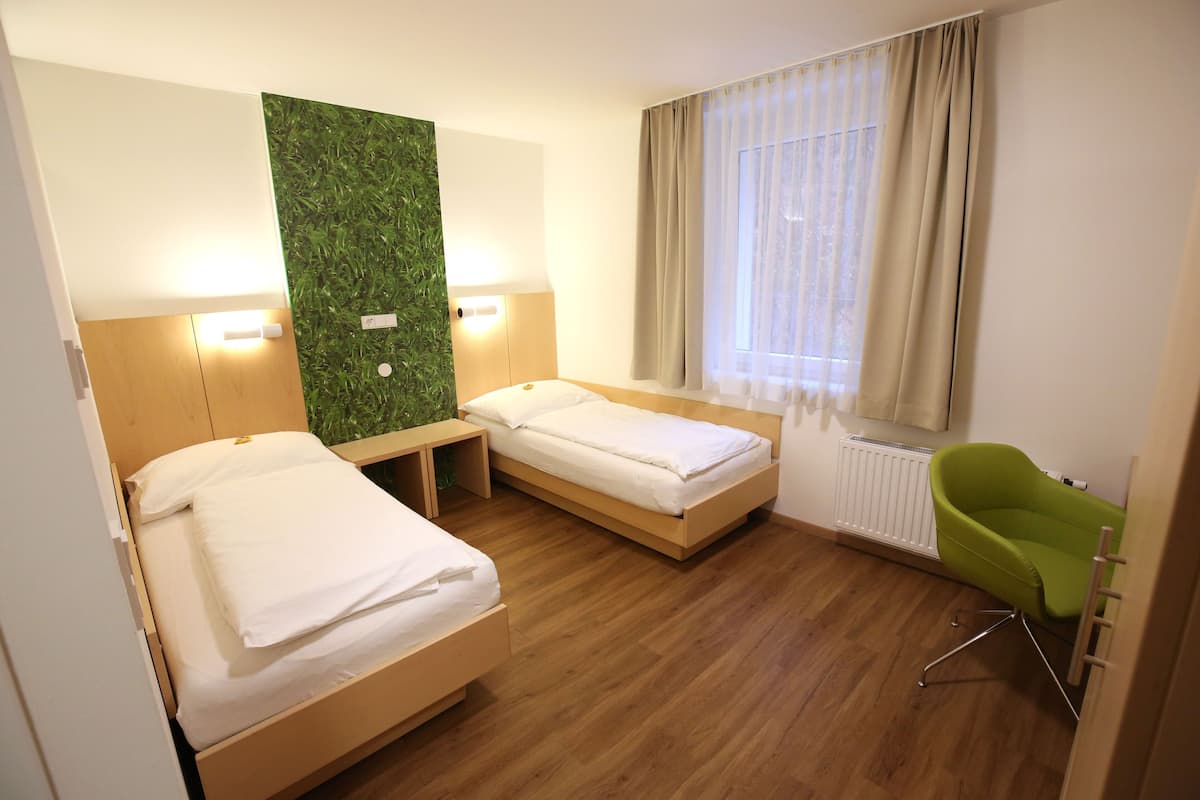 Habitación de hotel con dos camas, una pared verde, un radiador, una ventana con cortinas y una silla verde.