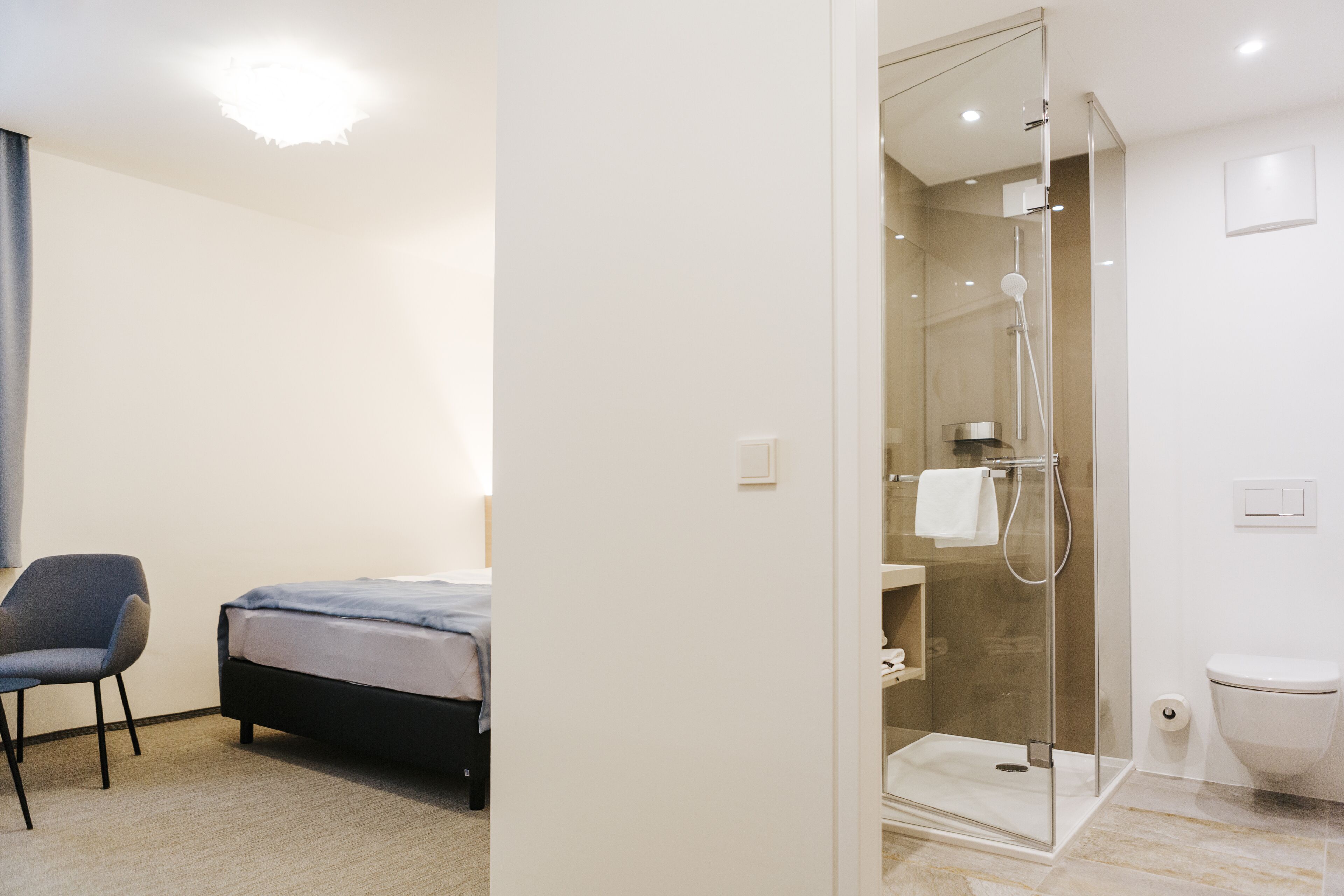 A modern hotel room with a bed, a chair, a glass-enclosed shower, and a toilet.