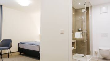 A modern hotel room with a bed, a chair, a glass-enclosed shower, and a toilet.