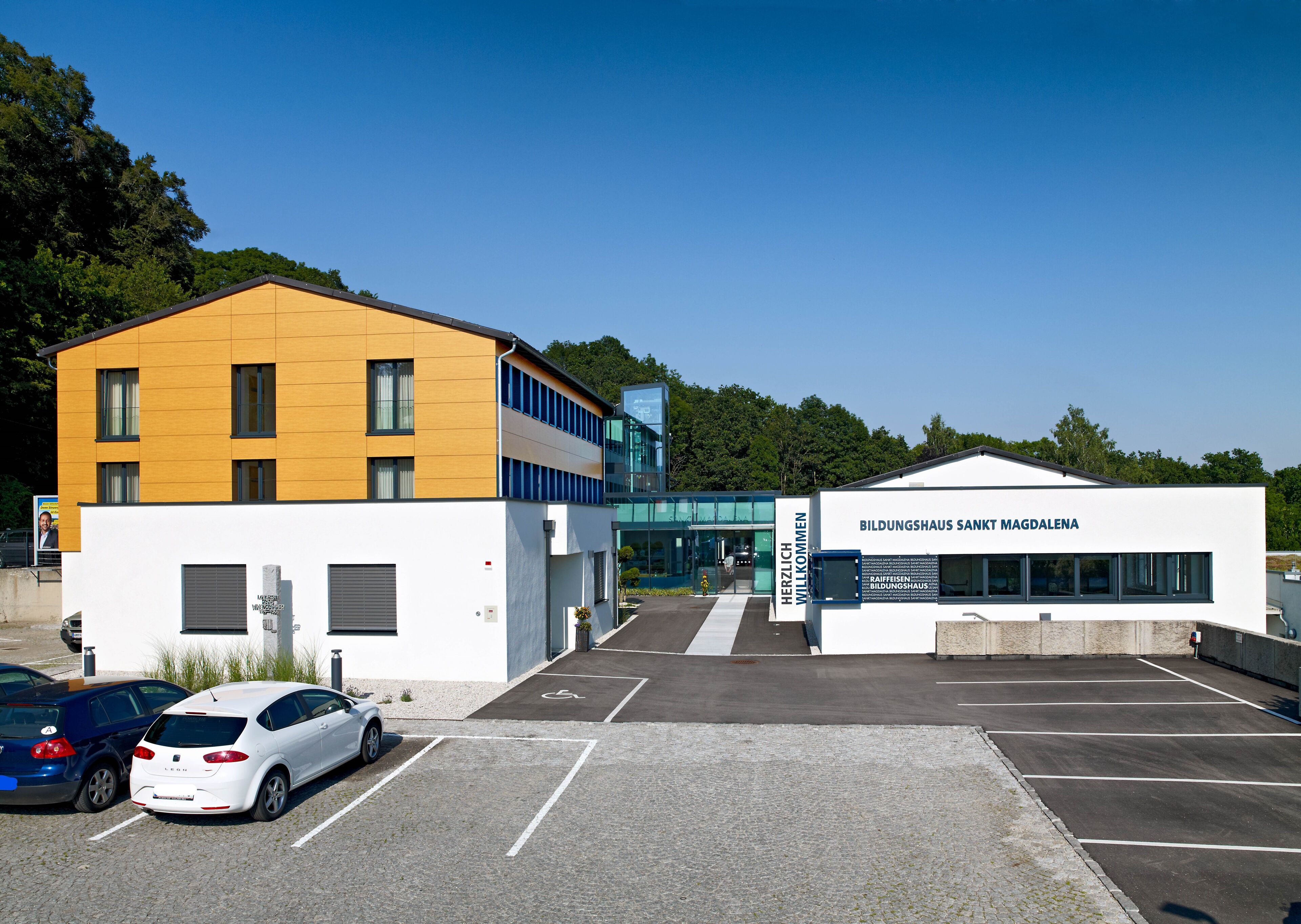 En modern byggnad med en gul och vit fasad, en parkeringsplats med markerade parkeringsrutor och en skylt som välkomnar besökare.