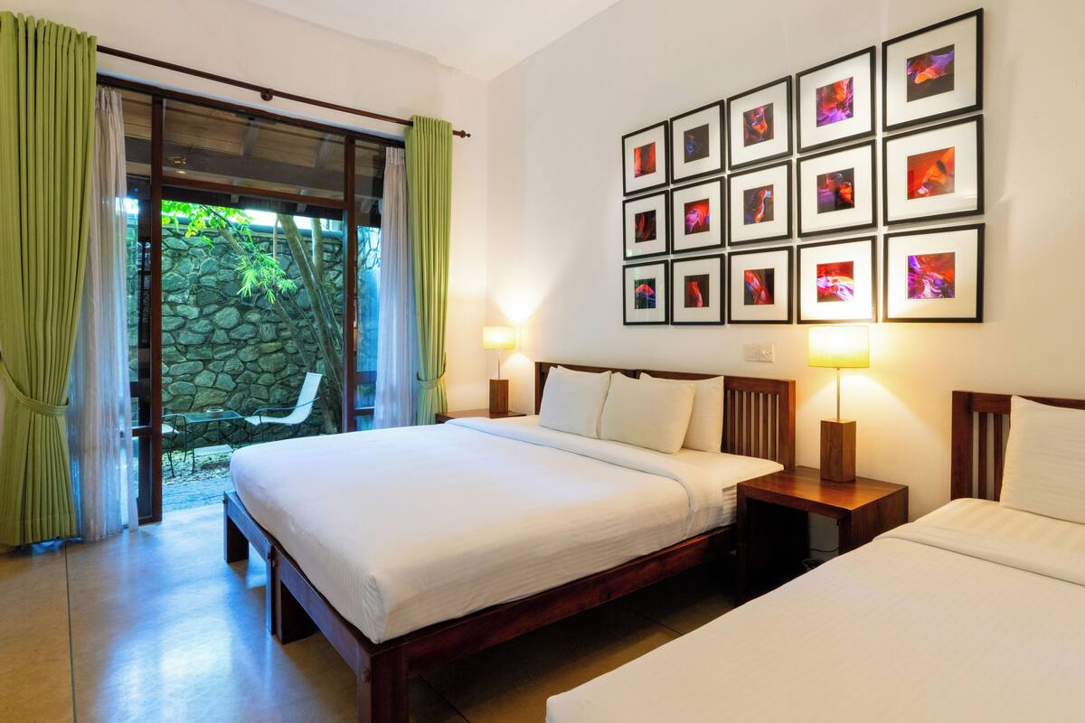 A hotel room with a large bed, two bedside tables, a wall with framed pictures, and a view of a stone wall and greenery outside.