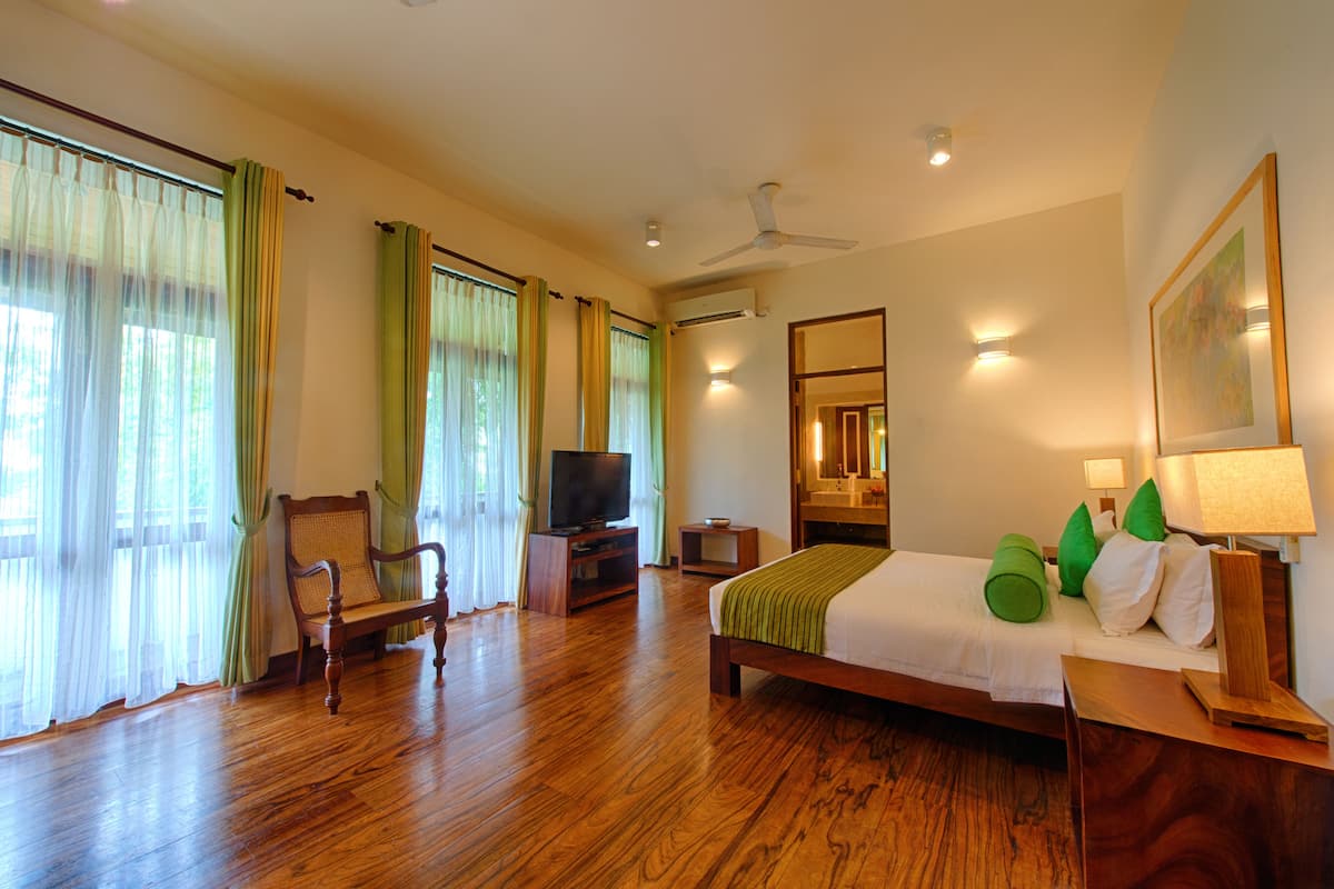 A hotel room with a wooden floor, a bed with green pillows, a television, a chair, a desk, and a mirror.