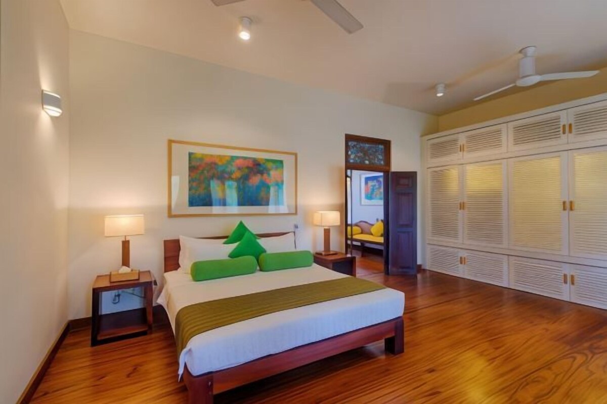 A hotel room with a wooden floor, a bed with green pillows, two bedside lamps, a painting on the wall, and a wardrobe with louvered doors.