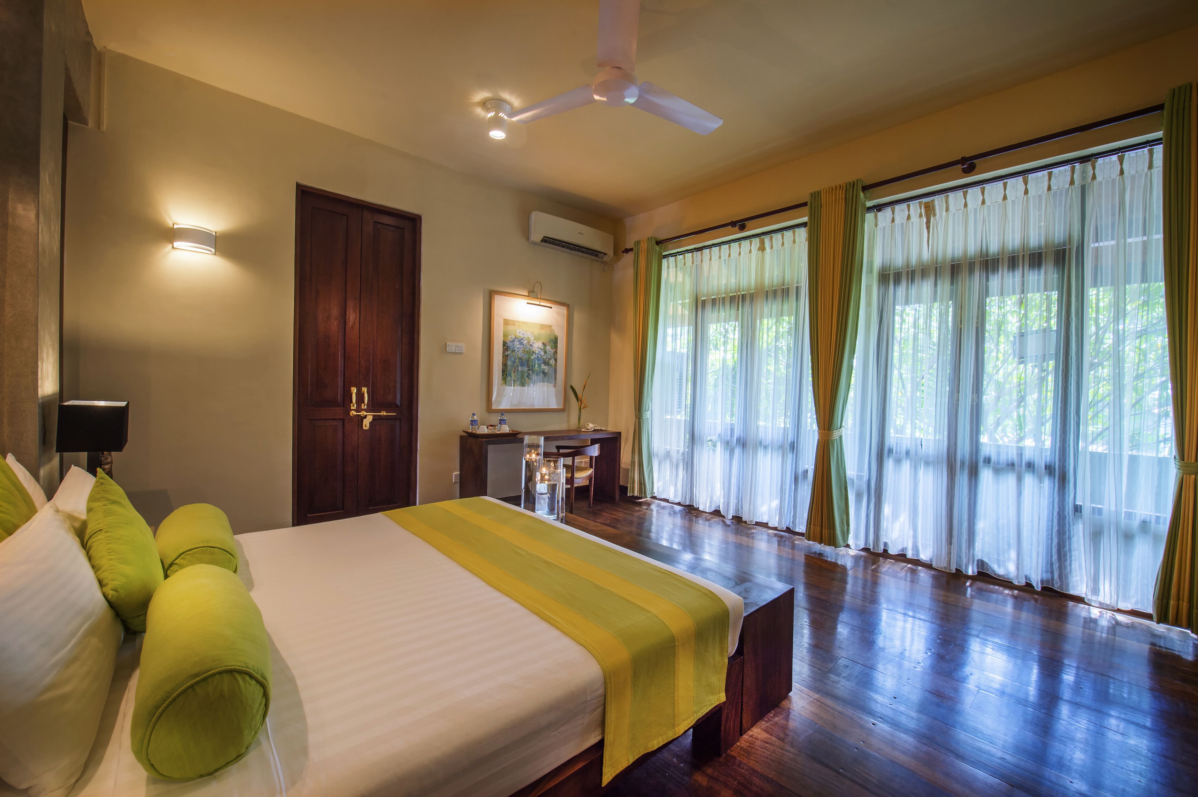 Habitación de hotel con cama, escritorio y una silla. Cuenta con ventilador de techo, ventana con cortinas y aire acondicionado de pared.