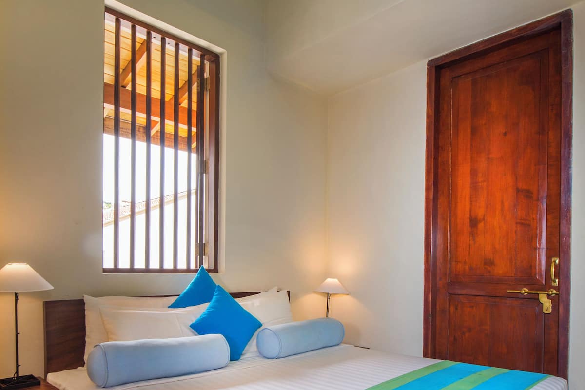 A hotel room with a bed, a window with bars, a wooden door, and a nightstand with a lamp.