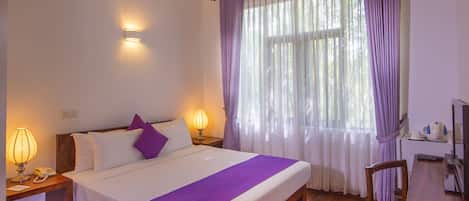 A hotel room with a bed, wooden floor, ceiling fan, bedside lamps, and a window with curtains.