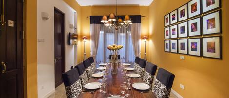 A dining room with a long table set for six, flanked by patterned chairs, a chandelier, and framed pictures on the wall.