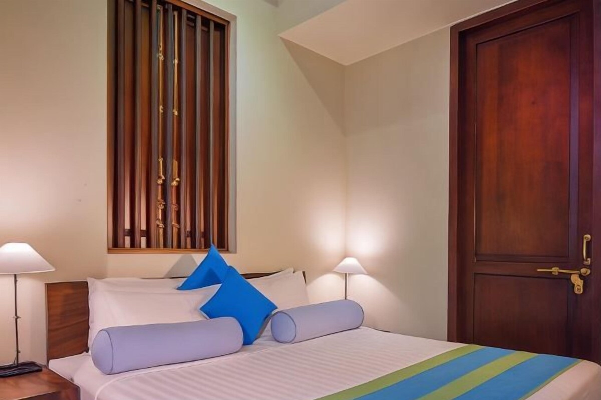 A hotel room with a bed, two bedside lamps, a wooden door, and a window with vertical blinds.