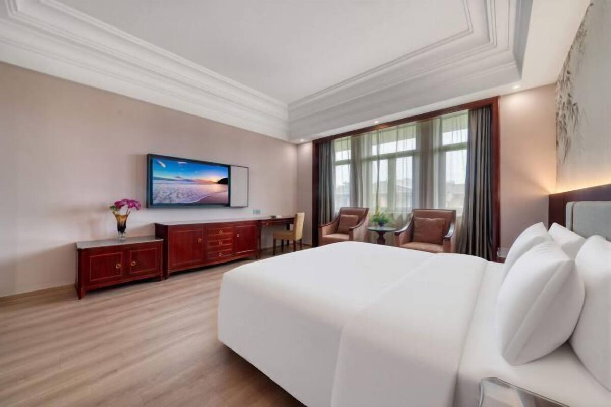 Quarto de hotel com uma cama grande, televisão na parede, escrivaninha com cadeira e janela com cortinas.