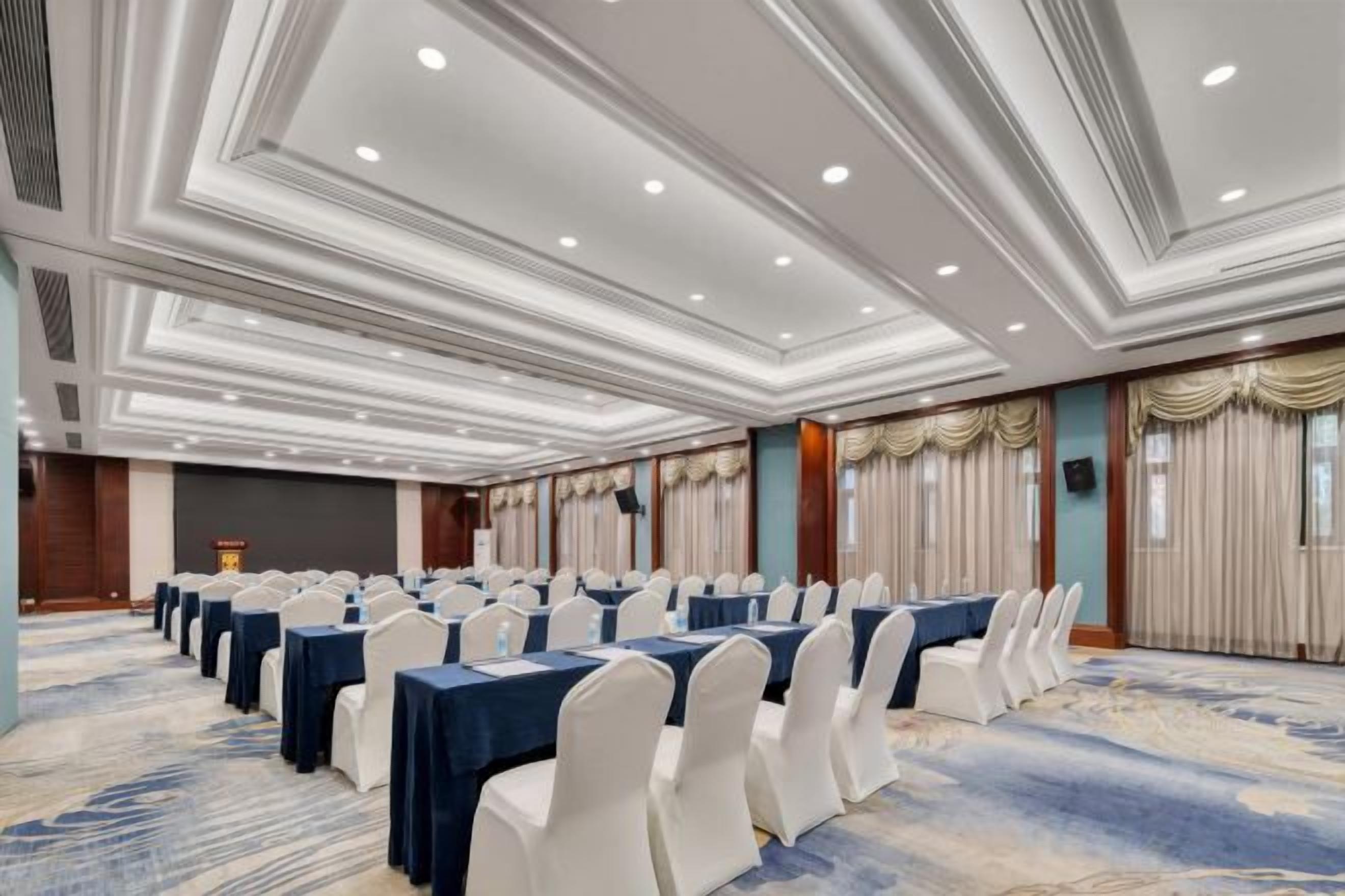 Un salón de conferencias con filas de mesas y sillas, piso alfombrado y un techo decorativo.