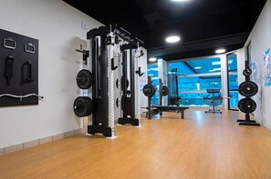 Un gimnasio con equipo de levantamiento de pesas, una banca y un estante fijado a la pared.
