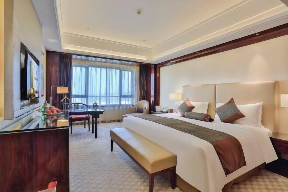 Une chambre d’hôtel avec un grand lit, un bureau avec une chaise, un banc et une grande fenêtre avec des rideaux.