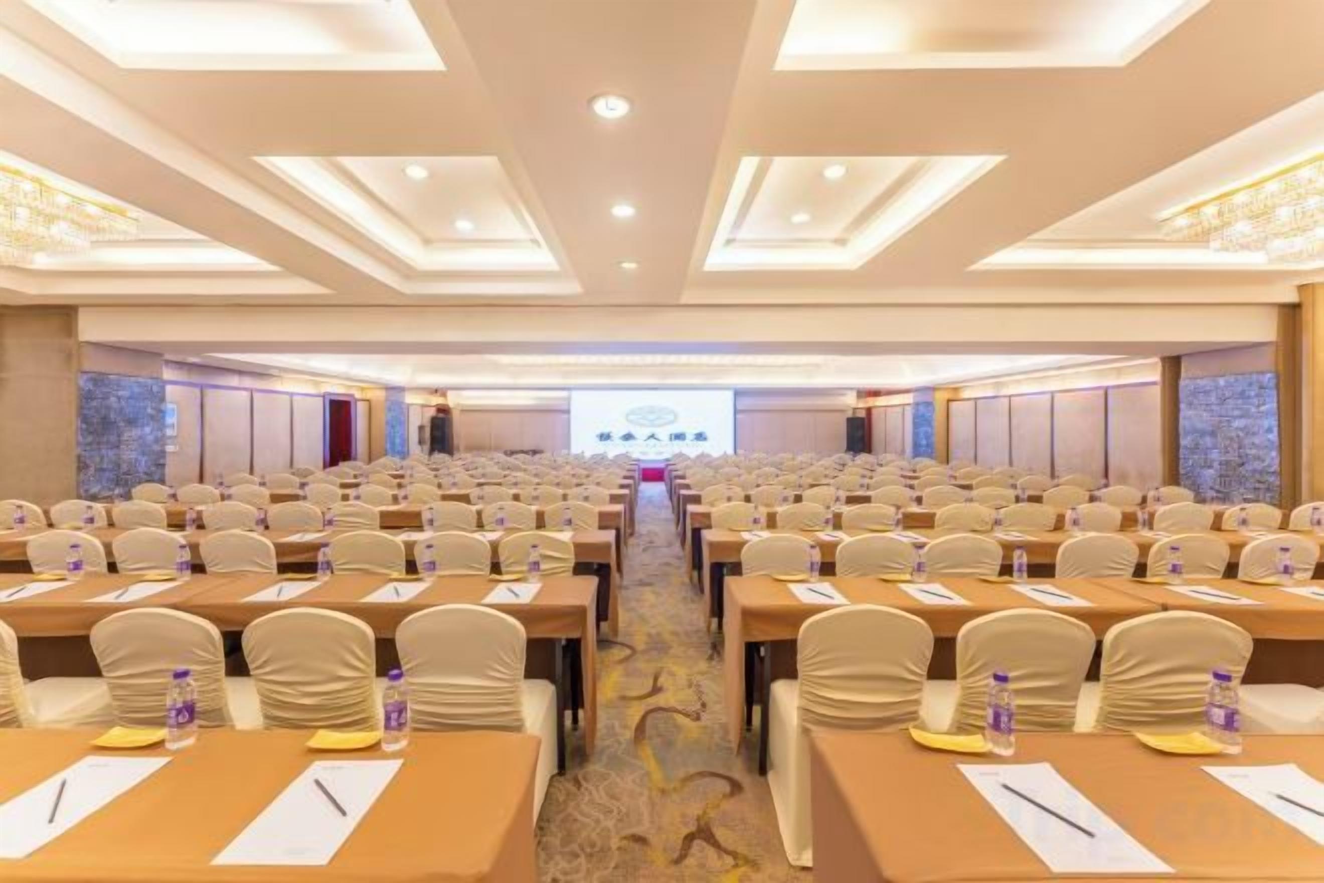 A conference room with rows of tables and chairs, a stage with a presentation screen, and a carpeted floor.