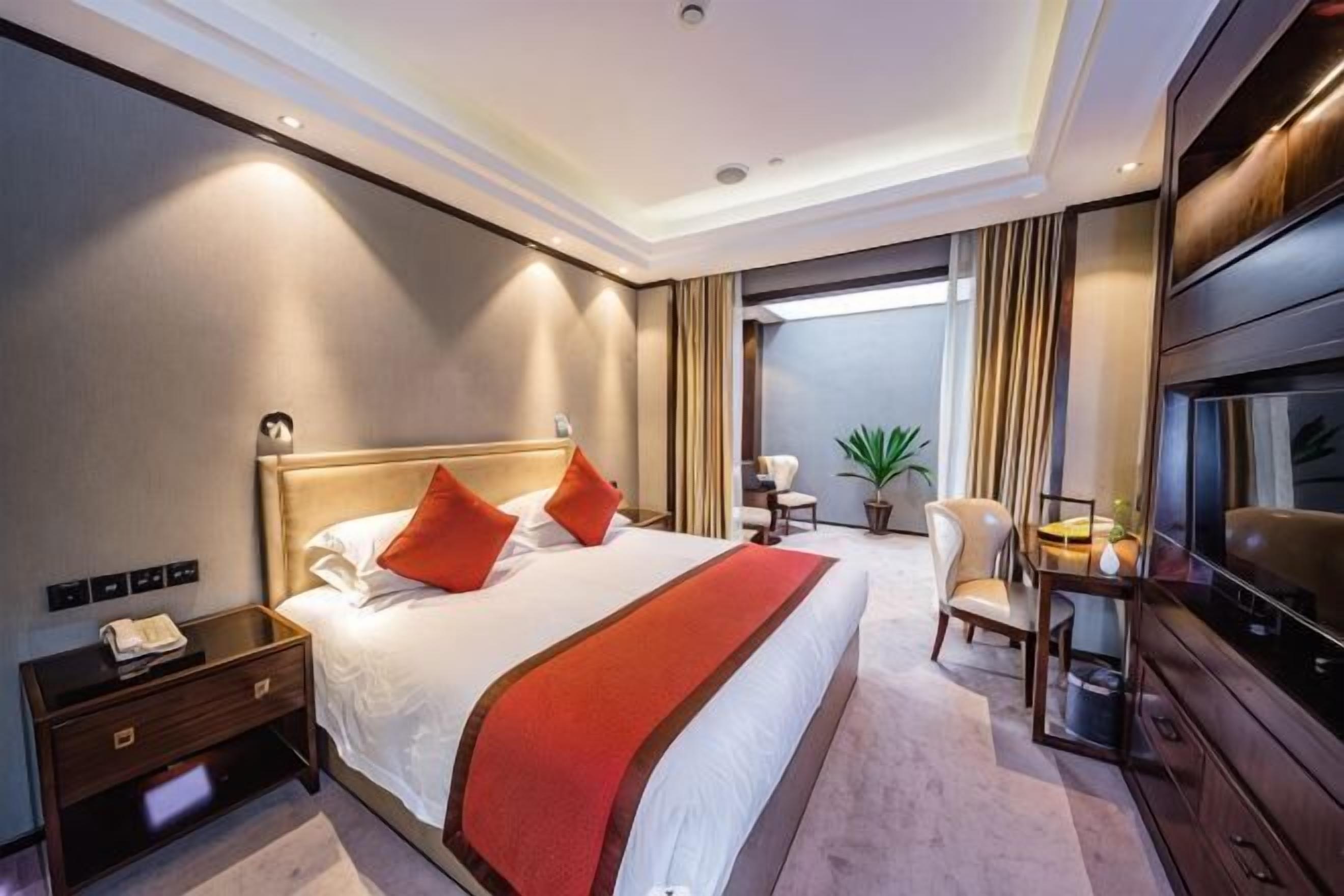 A hotel room with a large bed, a TV, a desk with a chair, and a plant.