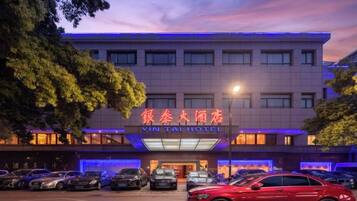 Un hotel con un letrero en un idioma extranjero, varios coches estacionados al frente, y un auto rojo en primer plano.