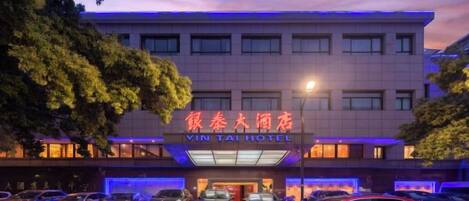 A hotel with a sign in a foreign language, multiple cars parked in front, and a red car in the foreground.