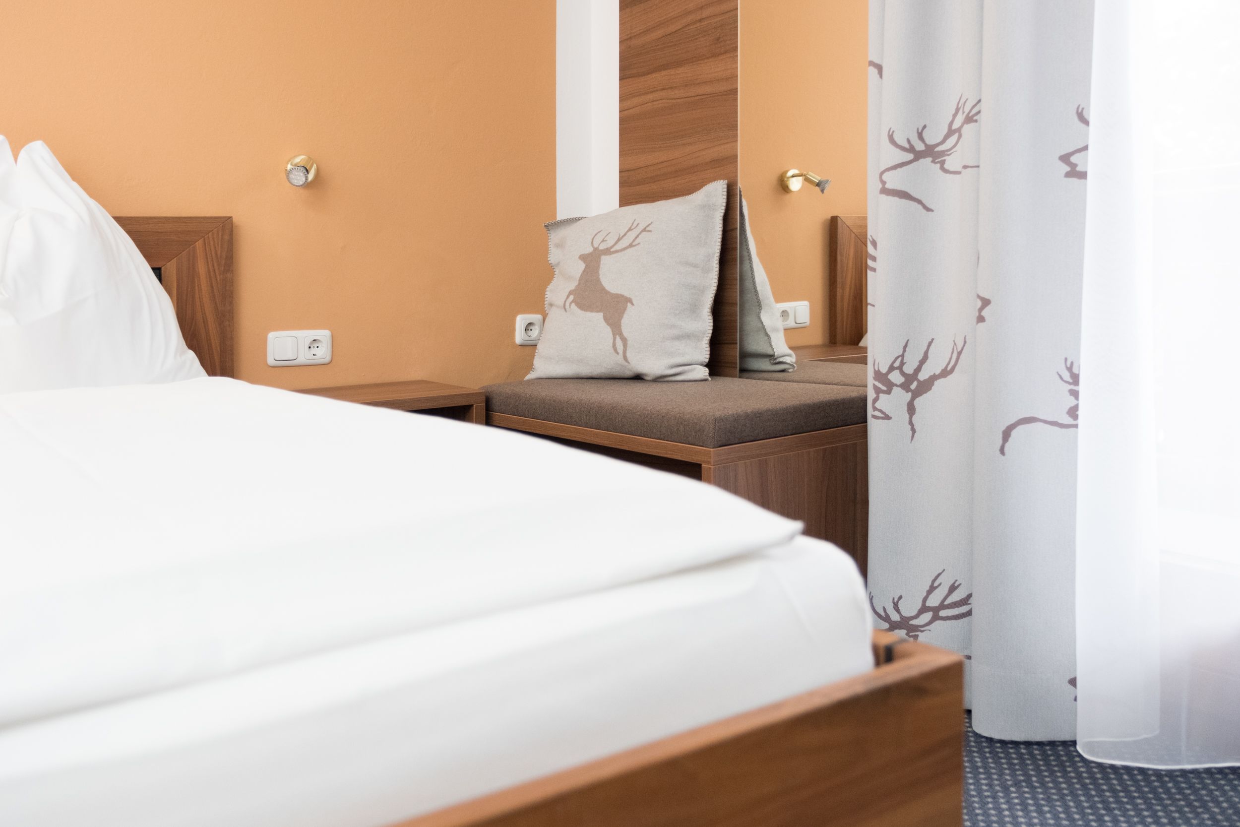 A hotel room with a bed, a nightstand, a lamp, a pillow with a deer design, and a curtain with a tree pattern.
