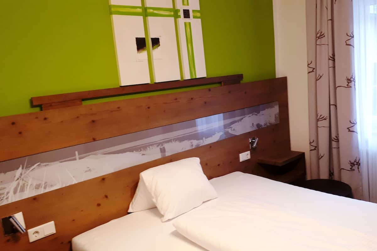 Habitación de hotel con cabecera de madera, cama con ropa de cama blanca, mesita de noche y pared verde con ventana y cortinas.