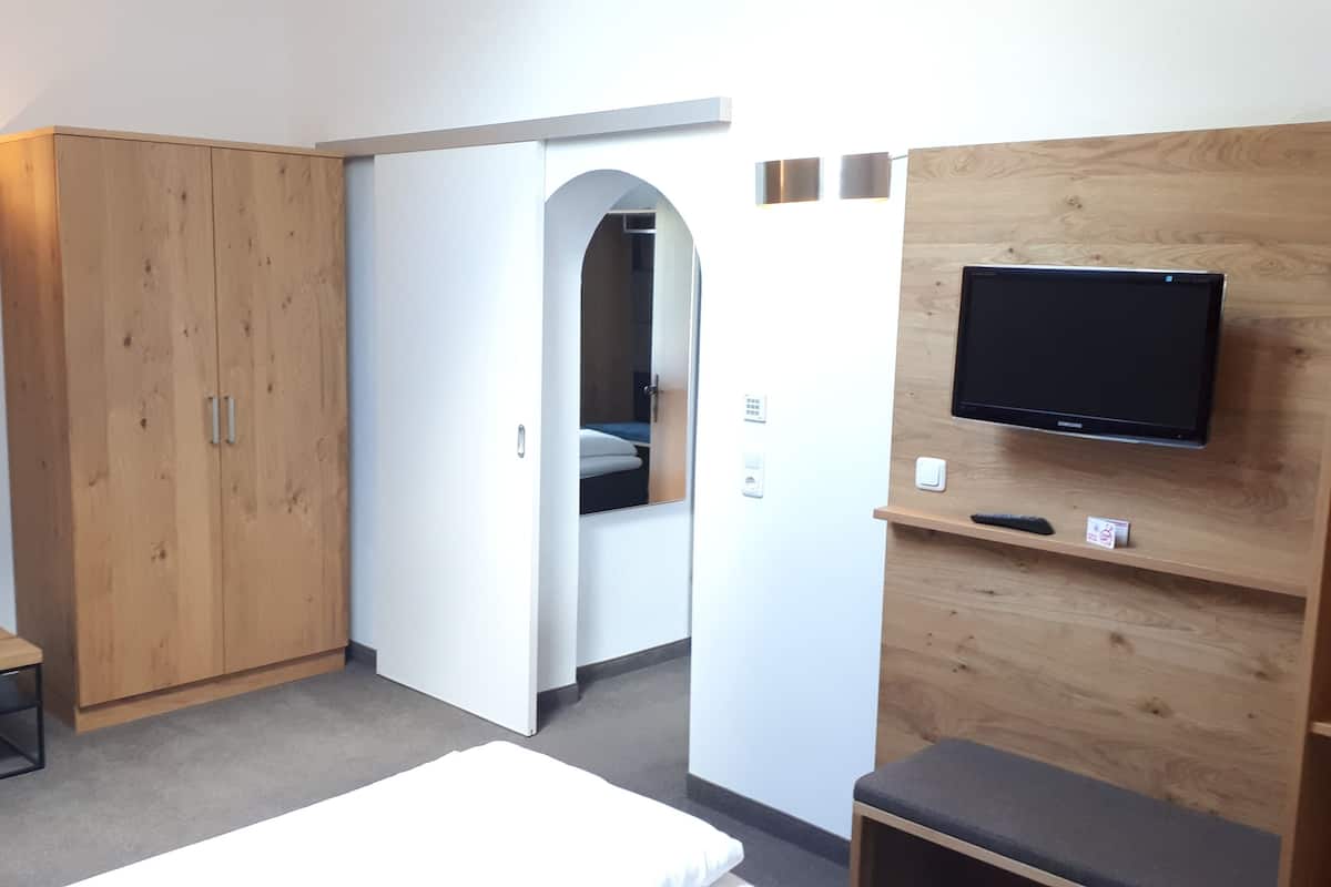 Habitación de hotel con armario, televisión fijada a un panel de madera, una mesita pequeña y una cama.