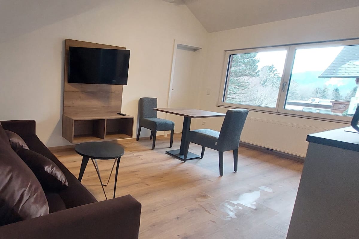 Una sala moderna con un sofá, una mesita, dos sillas, un televisor montado en un panel de madera y un amplio ventanal con vistas al exterior.