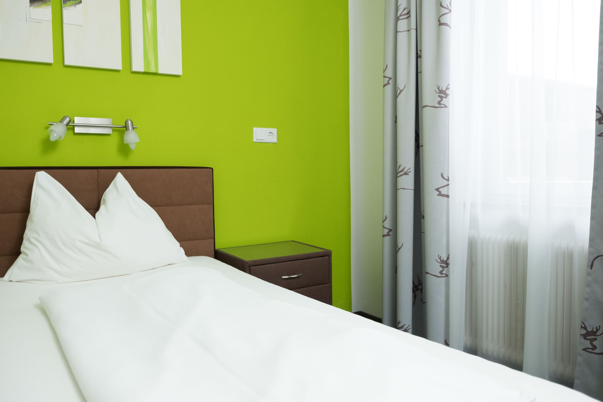 Ein Bett mit weißer Bettwäsche, ein braunes Kopfteil, ein Nachttisch mit Schublade, eine Wandleuchte und ein Fenster mit durchsichtigen Vorhängen.