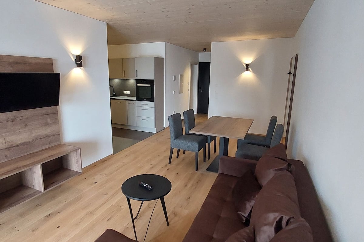 Una sala moderna con una pared de acento de madera, un televisor de pantalla plana, un comedor con mesa y sillas, y una cocina al fondo.