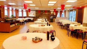 Un salón de banquetes con mesas redondas, algunas con sillas rojas, y una zona de bufé con mostrador y estanterías.
